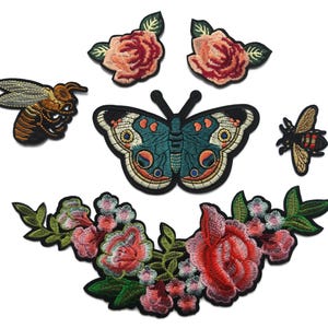 Puede incluir: Una colección de parches bordados que incluye una mariposa central grande con alas de color verde azulado y naranja, dos rosas individuales, dos abejas de diferentes tamaños y un amplio arreglo floral con rosas rojas y hojas verdes sobre fondo blanco.