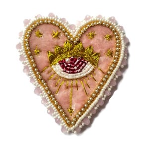 Op de afbeelding: Een hartvormige geborduurde patch met een roze fluwelen basis. Het ontwerp heeft een gouden kroon, een oog met rode en witte details en gouden stralen. De patch is omrand met witte en roze kralen en gouden draad.