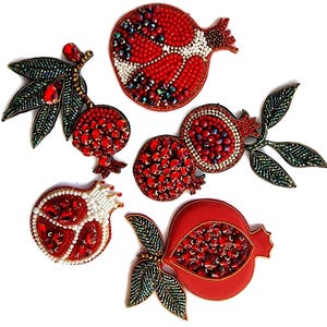 Könnte beinhalten: Eine Sammlung von aufwendig bestickten Granatapfel-Designs. Die Früchte sind hauptsächlich rot, mit weißen, grünen und schillernden Perlenakzenten. Einige sind ganz, andere aufgeschnitten, wodurch die juwelenfarbenen Samen sichtbar werden.