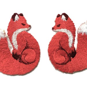 May include: Two embroidered fox patches. Each fox is curled up, with red fur, white accents, and a white-tipped tail. The patches are detailed with dark brown shading and are set against a white background.