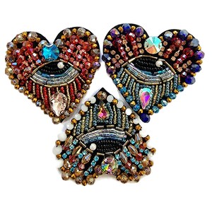 May include: Three heart-shaped beaded brooches with an eye design. The brooches feature a variety of colourful beads, including red, blue, gold, and iridescent gems. The eye design is centered on a black background.