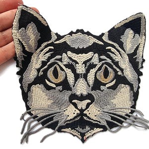 May include: Embroidered cat face patch with intricate detailing. The design features a black base with grey and beige highlights, including the eyes, nose, and whiskers. The patch is held by a hand.