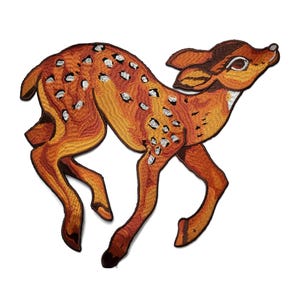 May include: Embroidered patch of a fawn in shades of orange and brown with white spots. The patch depicts the deer in a running pose, with its head and ears raised. The patch is likely for clothing or accessories.
