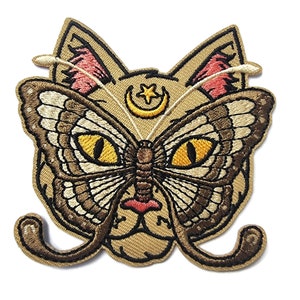 Puede incluir: Parche bordado termoadhesivo con un rostro de gato con alas de mariposa y una luna creciente y una estrella sobre la cabeza. El parche es beige con detalles marrones, amarillos y rosas.