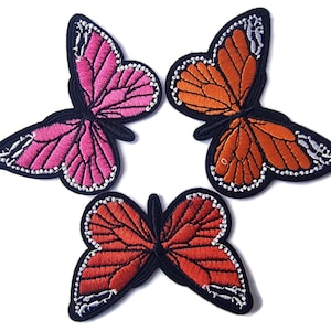 May include: Three embroidered iron-on patches featuring butterfly designs. The butterflies are pink, orange, and red with black outlines and white accents.