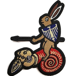May include: An embroidered patch featuring a brown rabbit standing on a red spiral base. The rabbit holds a horizontal spear and carries a blue shield with a white eagle emblem. A tan skull with dark details is positioned at the base of the spiral.