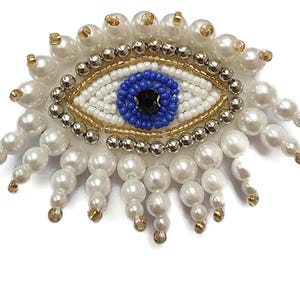 Può includere: Un ciondolo di occhio turco con perline, con un occhio blu e perline bianche e dorate. Il ciondolo è circondato da perline di perle bianche.