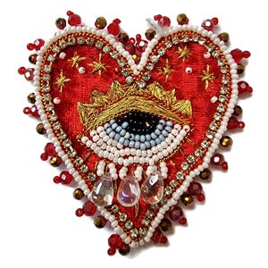 May include: A heart-shaped, red fabric brooch with an embroidered eye design. The brooch is embellished with white, red, and gold beads, rhinestones, and teardrop-shaped crystals. The eye has a black pupil and blue and white iris.
