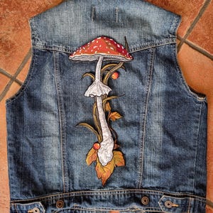 May include: A blue denim vest featuring an embroidered mushroom design on the back. The mushroom has a red cap with white spots, a white stem, and is surrounded by green and brown leaves.