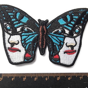 Puede incluir: Parche bordado para planchar de una mariposa azul y negra con dos caras. La mariposa tiene un cuerpo marrón y las caras tienen labios rojos.
