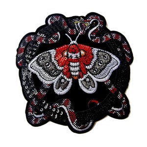 May include: Embroidered patch with a moth design, featuring red and white details, surrounded by a snake with red and white patterns. The patch is on a black background with a scalloped edge, enhanced with sequins.