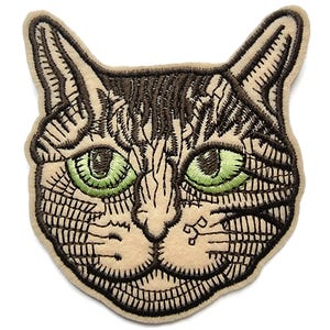 May include: Embroidered patch of a cat's face with brown and green fur. The cat has green eyes and a mischievous expression.