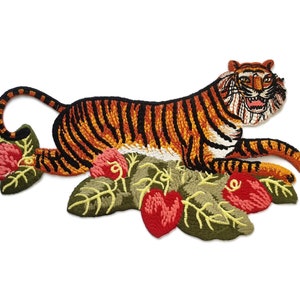 May include: An embroidered iron-on patch featuring a tiger with orange and black stripes, lying on a bed of green leaves with red flowers.