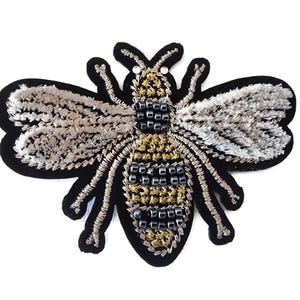 May include: A decorative bee-shaped patch, featuring intricate embroidery in black, silver, and gold. The wings are silver, the body is black and gold, and the patch has a black felt backing. The bee has two small, clear bead eyes.