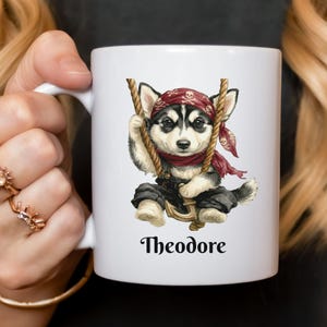 May include: White ceramic mug featuring a pirate-themed husky puppy illustration. The puppy wears a red bandana with skull and crossbones, black pants, and holds a rope. The name "Theodore" is printed below the image.
