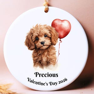 Pode incluir: Enfeite redondo de cerâmica branca com uma ilustração em aquarela de um cachorrinho marrom segurando um balão vermelho em forma de coração. O texto "Precious Valentine's Day 2026" está impresso abaixo. Uma corda dourada está presa para pendurar.