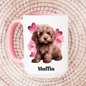 Pode incluir: Caneca de cerâmica branca com alça e borda rosa. A caneca apresenta uma ilustração de filhote marrom com corações rosa e o nome "Muffin" em texto preto. A caneca é projetada para bebidas.