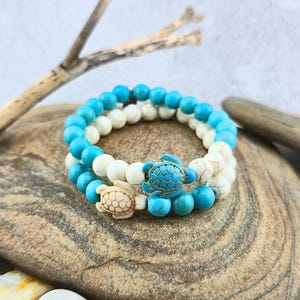 May include: Two beaded bracelets with turtle charms. One bracelet features turquoise and white beads, while the other has white beads. The turtle charms are in matching colors. The bracelets are displayed on a stone surface.