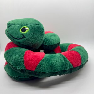 May include: A plush snake toy in shades of green and red. The snake's body is a vibrant green with red stripes. The head is green with a smiling face and yellow and black eyes. The toy is soft and cuddly, perfect for children.