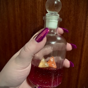 May include: A clear glass bottle with a round stopper, filled with a pink liquid and glitter. Two four-sided dice are inside the bottle.