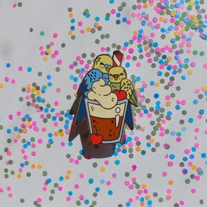 May include: A gold-tone enamel pin featuring two cartoon birds perched on top of a brown and white milkshake with a cherry on top. The birds are yellow, blue, and green.