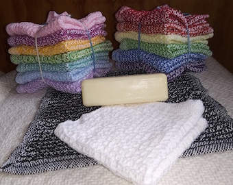 May include: A stack of colorful knitted dishcloths, a bar of soap, and two more dishcloths, one white and one black and white, on a white surface.