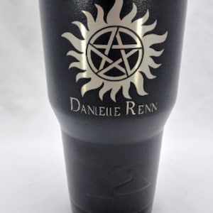 May include: A black 30oz tumbler with a silver lid. The tumbler has a silver  engraving of a pentagram with a sunburst around it. The text "DANIELLLE RENN" is engraved below the symbol.