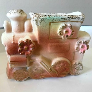 May include: A vintage ceramic train planter with pink flowers and gold accents. The planter is painted in shades of brown, pink, and green.