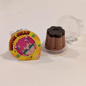 May include: A coffee jelly charm with a cartoon character illustration and the words "COFFEE JELLY". Next to it is a coffee jelly dessert in a clear plastic container. The dessert is brown with a dark brown top.