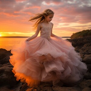 May include: A young girl in a pink tulle dress stands on a rocky beach at sunset. Her hair is blowing in the wind and the dress is flowing around her.