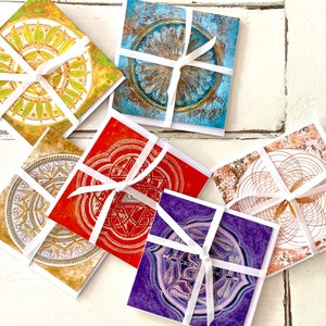 May include: Six colourful cards with intricate geometric designs and white ribbon tied around them. The cards are on a white wooden surface.