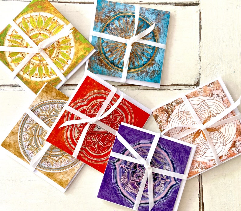 May include: Six colourful cards with intricate geometric designs and white ribbon tied around them. The cards are on a white wooden surface.