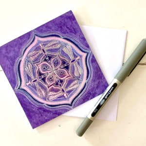 May include: Purple greeting card with a mandala design featuring pink dragonflies. The mandala is surrounded by a purple border.