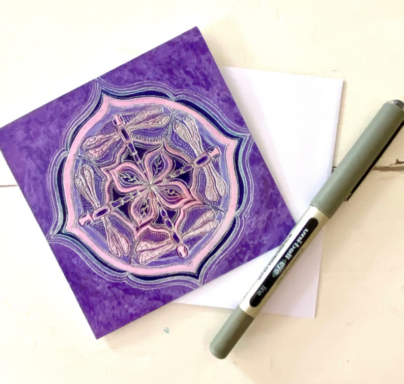 May include: Purple greeting card with a mandala design featuring pink dragonflies. The mandala is surrounded by a purple border.