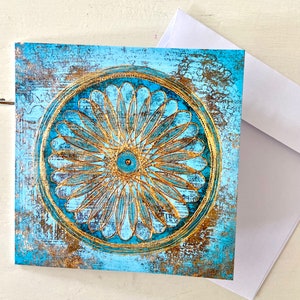 May include: A square card featuring a blue and gold mandala design. The mandala has a circular pattern with a central eye and radiating petals. The background is a distressed blue with gold accents.