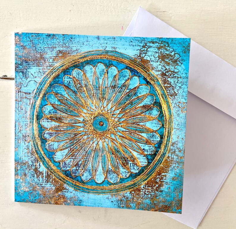 May include: A square card featuring a blue and gold mandala design. The mandala has a circular pattern with a central eye and radiating petals. The background is a distressed blue with gold accents.