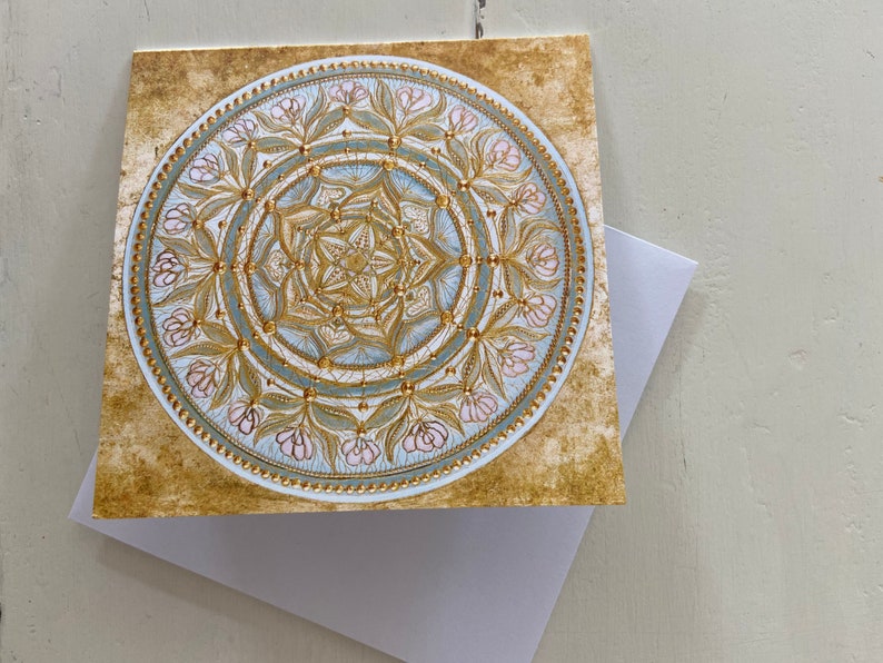 May include: A gold and blue mandala with pink floral accents on a textured gold background. The mandala is a circular design with intricate details and a geometric pattern.