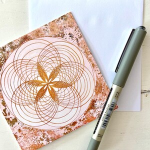 May include: A pink and gold card with a geometric design of overlapping circles and a flower in the centre. The card is on a white surface with a pen.