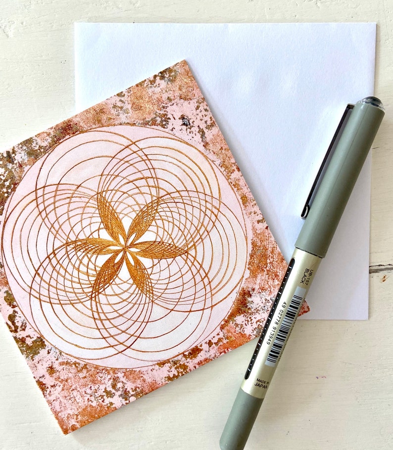 May include: A pink and gold card with a geometric design of overlapping circles and a flower in the centre. The card is on a white surface with a pen.