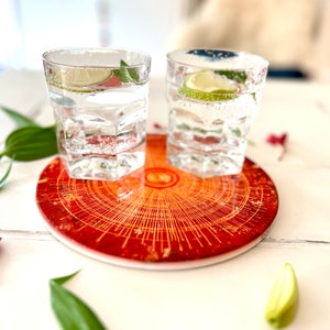 Könnte beinhalten: Zwei klare Glasbecher gefüllt mit Sprudelwasser und Limettenscheiben, die auf einem runden Untersetzer in Rot und Gold mit geometrischem Muster liegen.