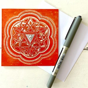 May include: A greeting card featuring a geometric design in silver on an orange background. The design is a stylised flower with a central triangle.