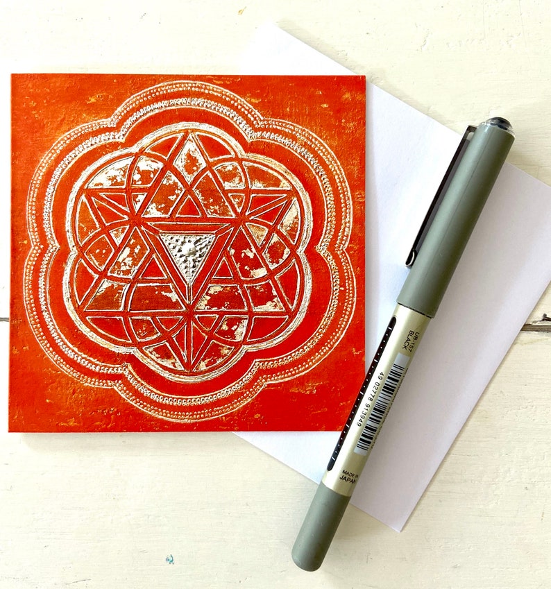 May include: A greeting card featuring a geometric design in silver on an orange background. The design is a stylised flower with a central triangle.