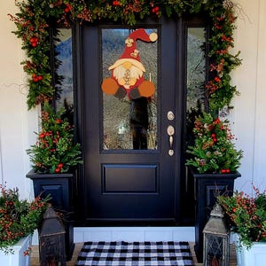 May include: A black front door with a red and white gnome door hanger. The door is decorated with a green garland with red berries. There are two white planters with green plants and red berries on either side of the door. A black and white checkered doormat is on the ground in front of the door.