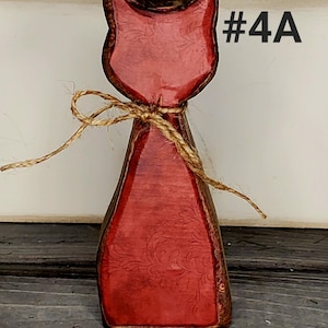 May include: Wooden cat figurine with a red finish and a decorative floral pattern. The cat is tied with twine and has the text "#4A" in the upper right corner. The figurine is on a wooden surface.
