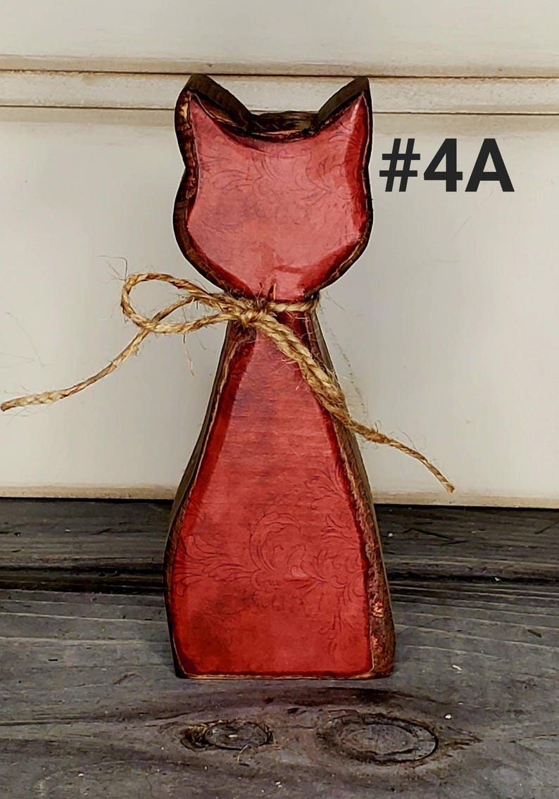 May include: Wooden cat figurine with a red finish and a decorative floral pattern. The cat is tied with twine and has the text "#4A" in the upper right corner. The figurine is on a wooden surface.
