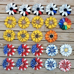 May include: A collection of 30 wooden flower-shaped wall decor pieces in various colors and patterns. The flowers are arranged in rows on a wooden surface. Some of the flowers have a patriotic theme with red, white, and blue colors. Others have a sunflower theme with yellow and black colors. There are also flowers with a black and white color scheme.