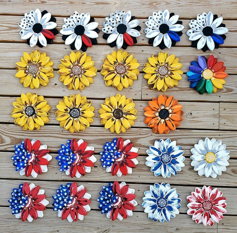 May include: A collection of 30 wooden flower-shaped wall decor pieces in various colors and patterns. The flowers are arranged in rows on a wooden surface. Some of the flowers have a patriotic theme with red, white, and blue colors. Others have a sunflower theme with yellow and black colors. There are also flowers with a black and white color scheme.