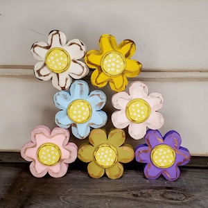 May include: Seven wooden flower cutouts in various colors: white, yellow, blue, pink, olive green, and purple. Each flower has a yellow center with white dots. The flowers are arranged on a wooden surface.