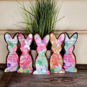 May include: Five wooden bunny cutouts with floral patterns in pink, green, orange, and blue. The bunnies stand in a row, with a brown outline, in front of a vase with tall green grass.
