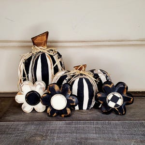 Może przedstawiać: Zestaw drewnianych dekoracji w kształcie dyni i kwiatów. Dynie są w czarno-białe paski z drewnianymi łodygami i akcentami z rafii. Kwiaty mają czarno-białe szachownice lub jednolite czarne środki.